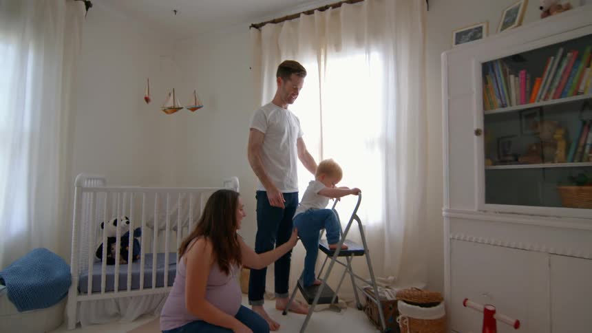 Parents help their toddler climb a small ladder in a softly lit nursery