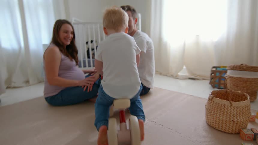 A young boy rides a toy bike as his smiling parents sit nearby in a bright nursery
