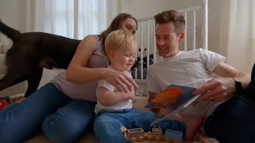 A pregnant mother and father sit on the floor with their child reading a book and having fun while their dog walks behind them in the childs room around midday