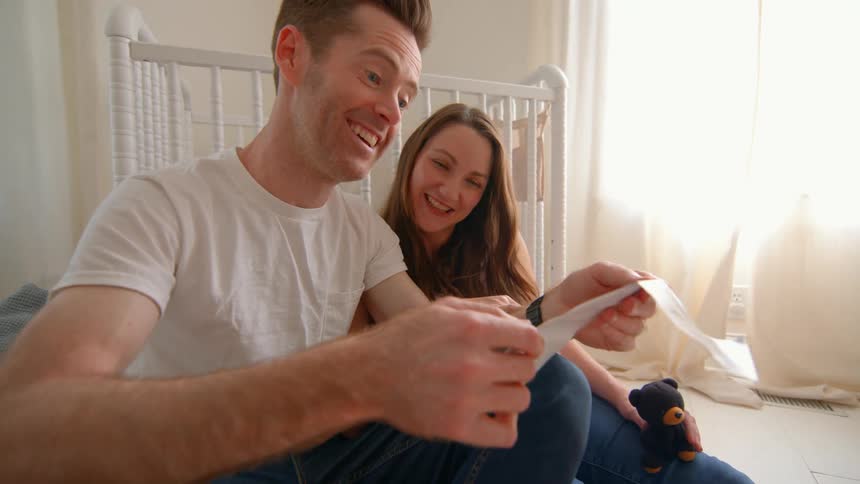A pregnant woman and a man sit on the floor in their baby room laughing as they look at their babys ultrasound scan