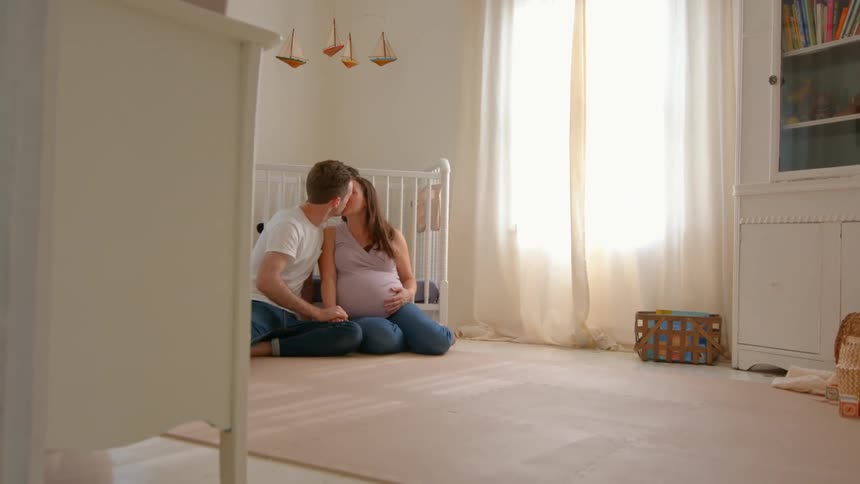 A couple sits on the floor in a baby room with a crib behind them kissing each other as the woman heavily pregnant rests beside her partner