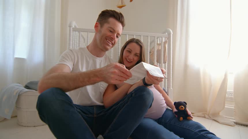 A pregnant woman and a man sit on the floor in their baby room smiling as they look at their babys ultrasound scan