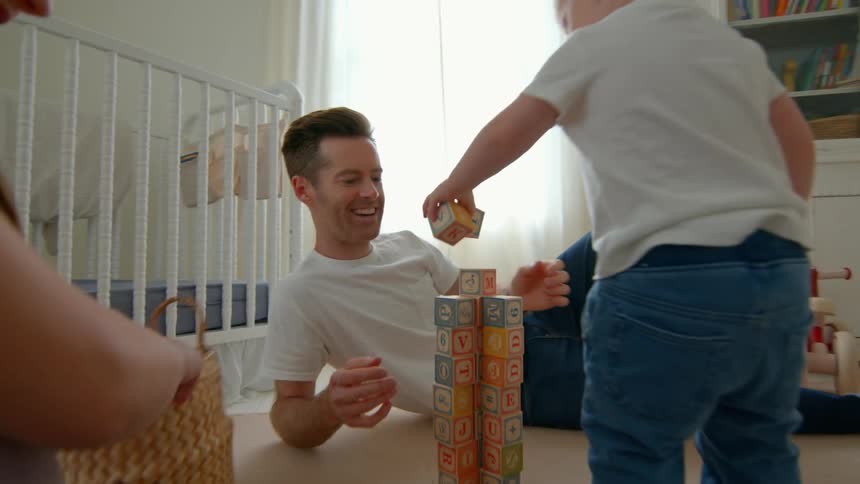 A pregnant mother and father sit on the floor with their toddler son picking up alphabet blocks and stacking them in the bedroom around midday as everyone laughs with excitement