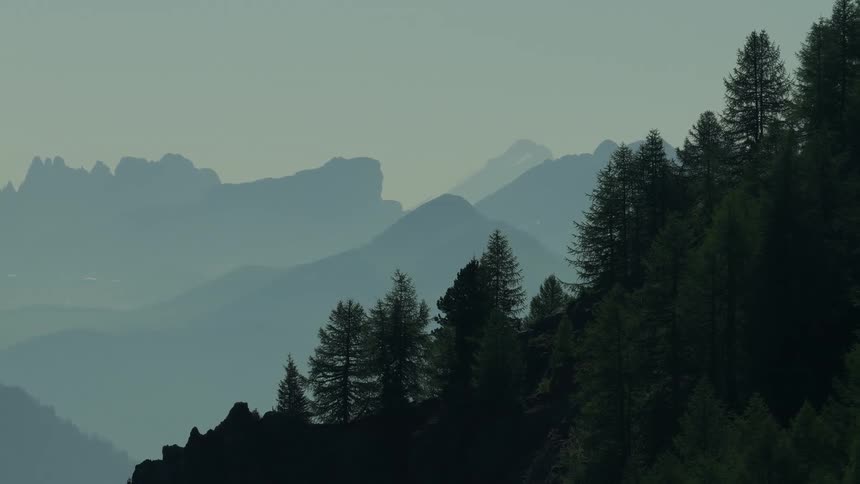 An aerial view looks over a dark pinecovered mountain revealing layers of hazy silhouetted mountain peaks in the distance