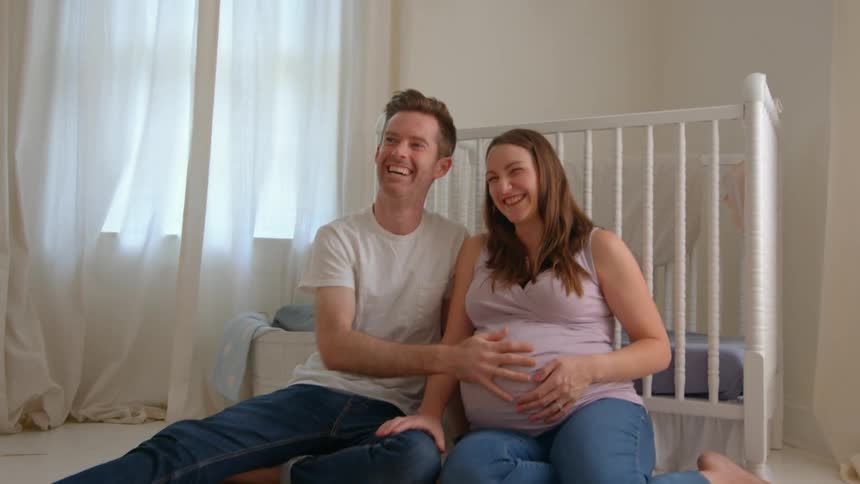 A happy couple sits on the nursery floor smiling and touching the womans pregnant belly