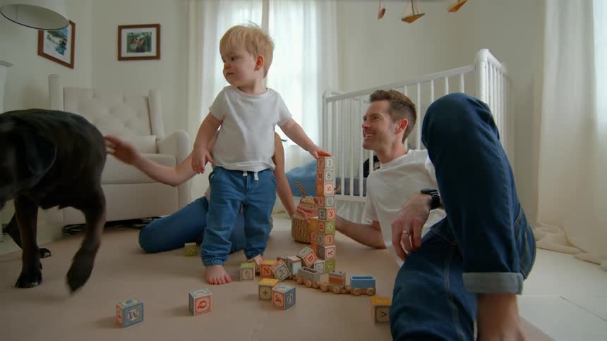 A father plays with his toddler son arranging alphabet blocks on the floor while the pregnant mother playfully knocks over the blocks with a bag causing them to fall they all start laughing and enjoy the moment as their pet dog roams around the room