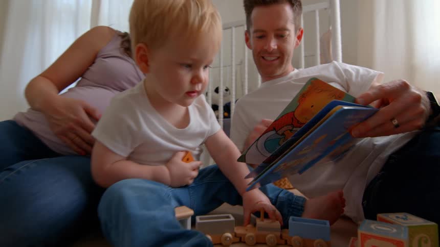 Pregnant mother with little son next to father reading a book in a room with toys
