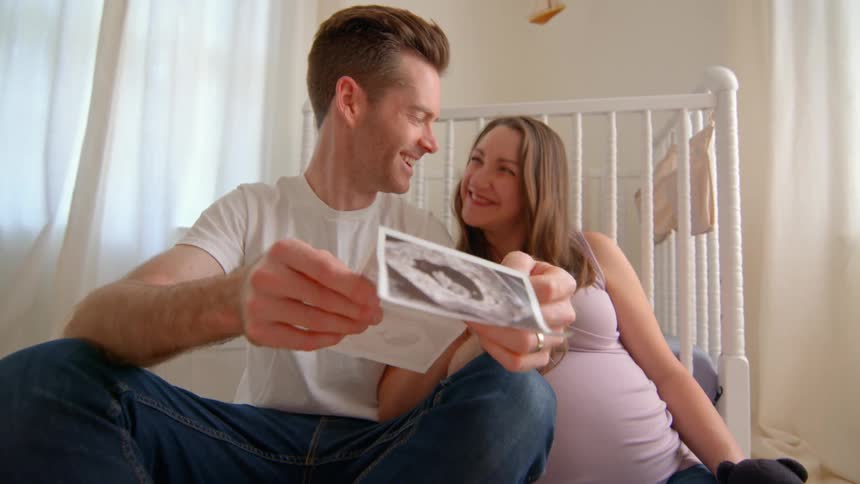 A couple sits on the nursery floor smiling at each other while holding an ultrasound photo