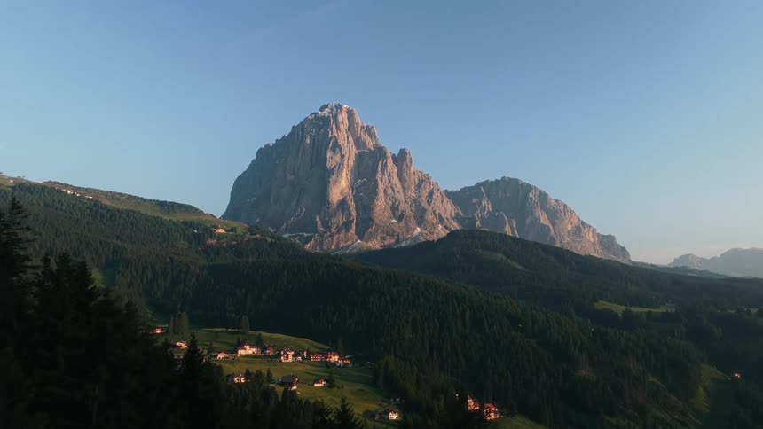 A majestic mountain range towers over a patchwork of cultivated fields and a scattered rural settlement in a sunlit valley