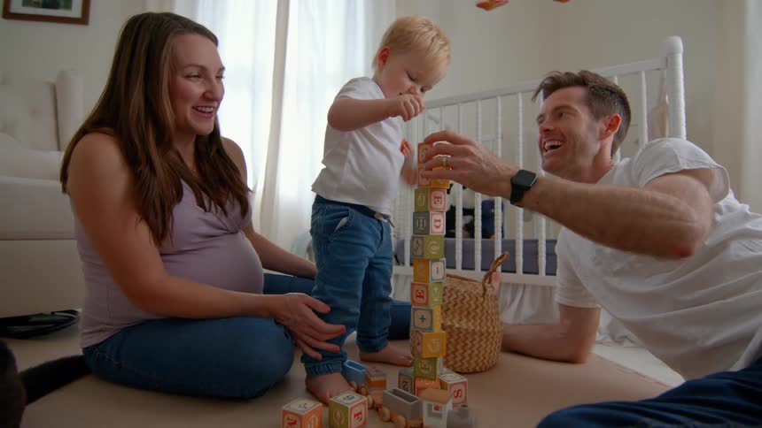 A family plays together with blocks on the nursery floor while a dog runs through the room