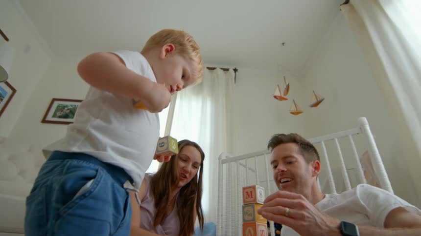A father plays with his toddler son arranging alphabet blocks on the floor while the pregnant mother watches from the side in a bedroom around midday