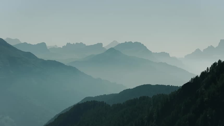 An aerial view flies forward over a vast mountain range revealing layers of hazy silhouetted peaks receding into the distance