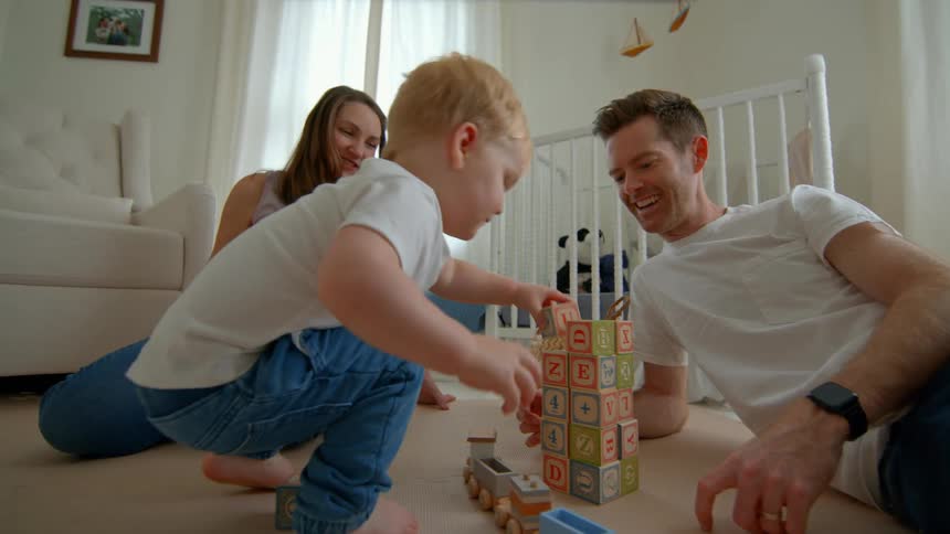 A child stacks colorful blocks on the floor while two adults watch attentively in a warmly lit nursery with a crib and hanging mobile