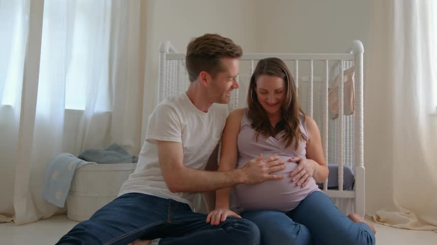 A couple sits on the floor of a softly lit nursery the man gently placing a hand on his partners pregnant belly near a white crib