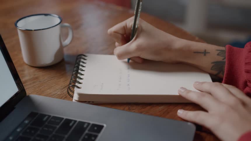 Hand with a pen writing on a notepad on a table next to an open laptop