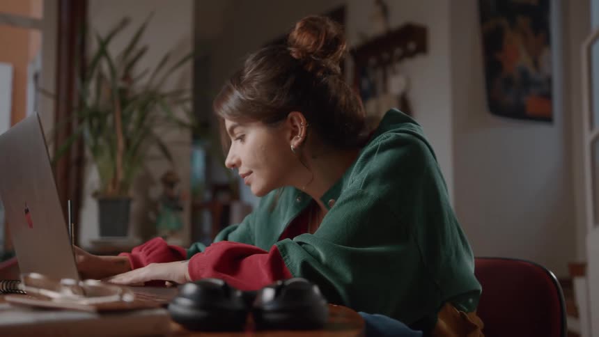 A woman is sitting at a desk using a laptop and writing in a notebook with headphones and a plant visible in the background