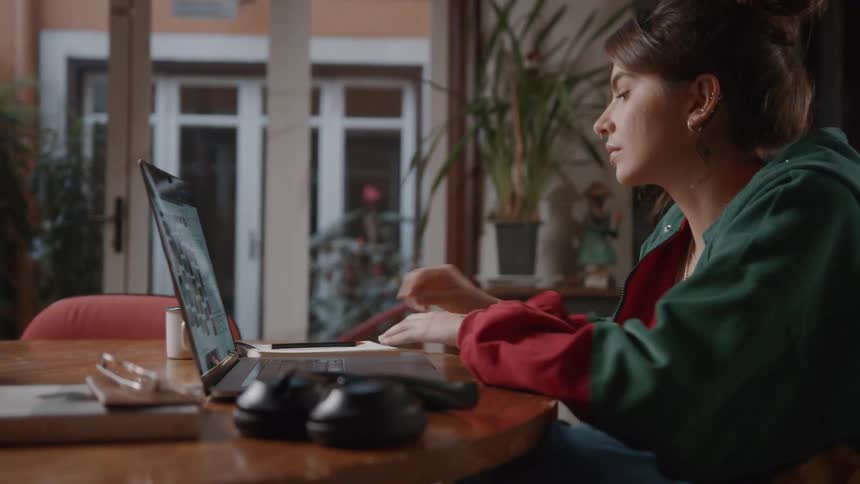 A young woman in a hoodie sits at a wooden table taking notes in a spiral notebook while looking at a photo gallery on her laptop