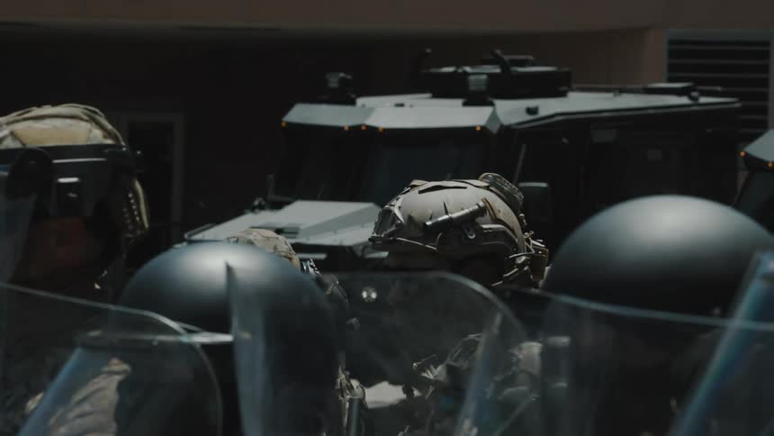 Law enforcement personnel in helmets and holding clear shields stand in front of a dark armored vehicle