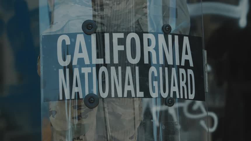 A clear shield labeled california national guard rests in front of a person in camouflage near a graffiticovered wall