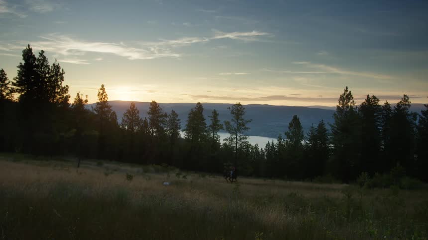 A person rides a horse past a grassland surrounded by trees with a lake visible in the distance at sunset