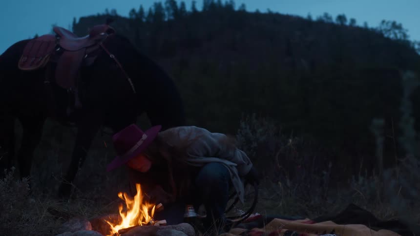 A woman wearing a cowboy hat starts a campfire in the woods in the evening with a horse standing behind her