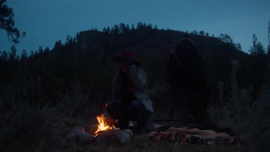 A woman sits by a campfire at dusk with her horse behind her in a quiet forest clearing