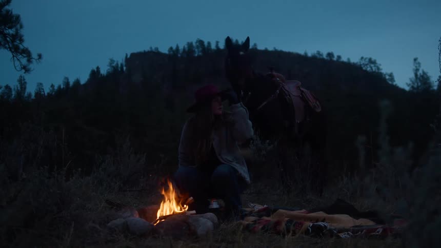 Woman in front of a bonfire in the woods at dusk next to a horse