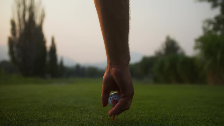 Close up of a man fixing a golf tee and ball on a field outdoors at sunset