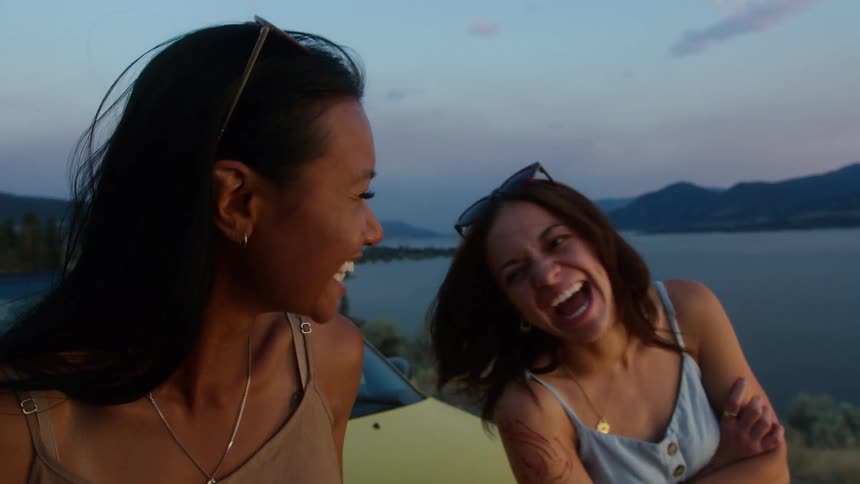 Two friends recline on a car near a lakeshore at sunset laughing and enjoying a carefree moment together