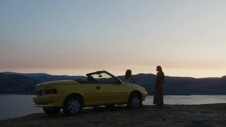 Two people stand near a yellow convertible at sunset overlooking a serene lake and hills