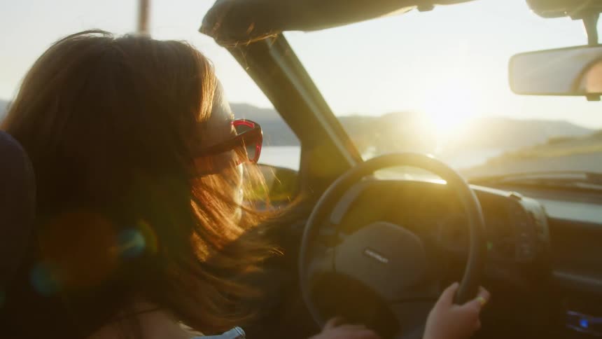 A young woman wearing sunglasses drives an openroof car around midday on a coastal road