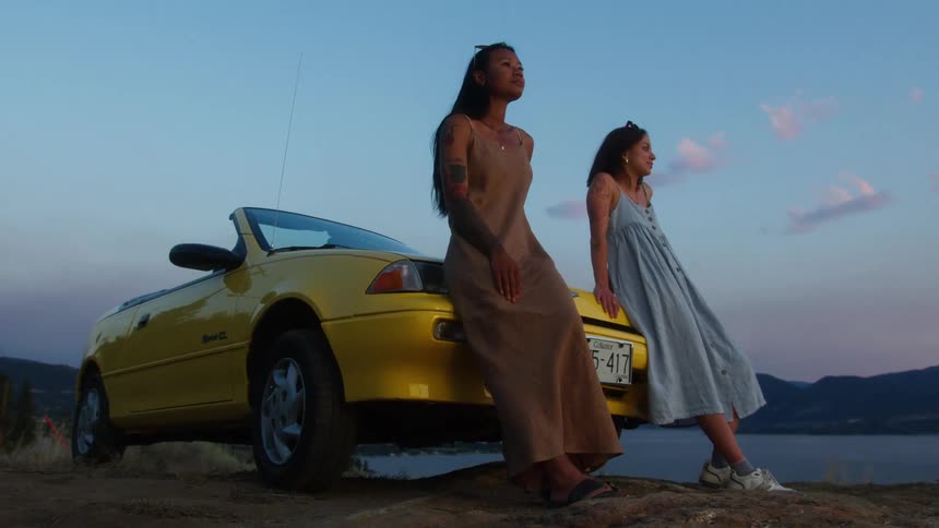 Two women lean on a car and stare into the distance