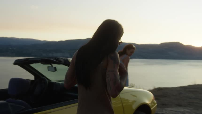 Two women got out of the car parked by the river and laughed while standing near the car bonnet at sunset