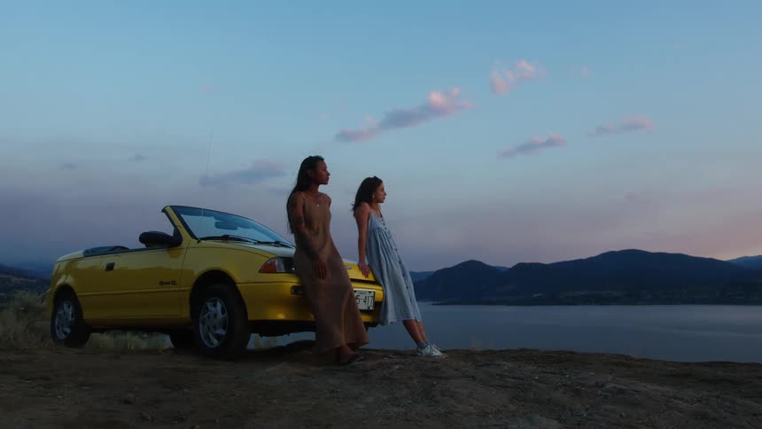 Two women stand beside a yellow convertible overlooking a lake at sunset