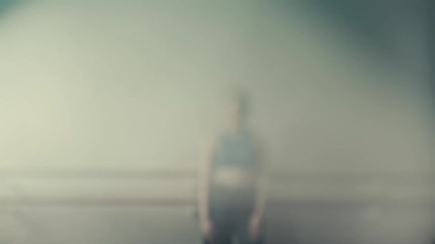 A young woman wearing a blue top stands beside a wall indoors around midday