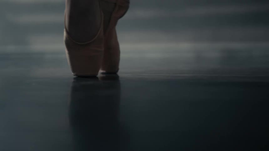 A ballerina balances en pointe in worn satin shoes her feet aligned on a reflective floor under soft studio lighting