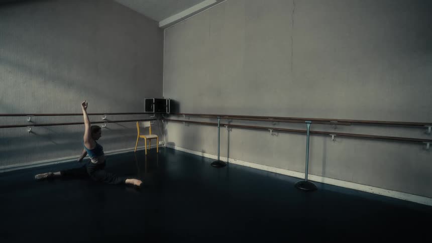 A woman is performing a split on the floor of a dance studio with ballet barres along the walls and a yellow chair in the corner