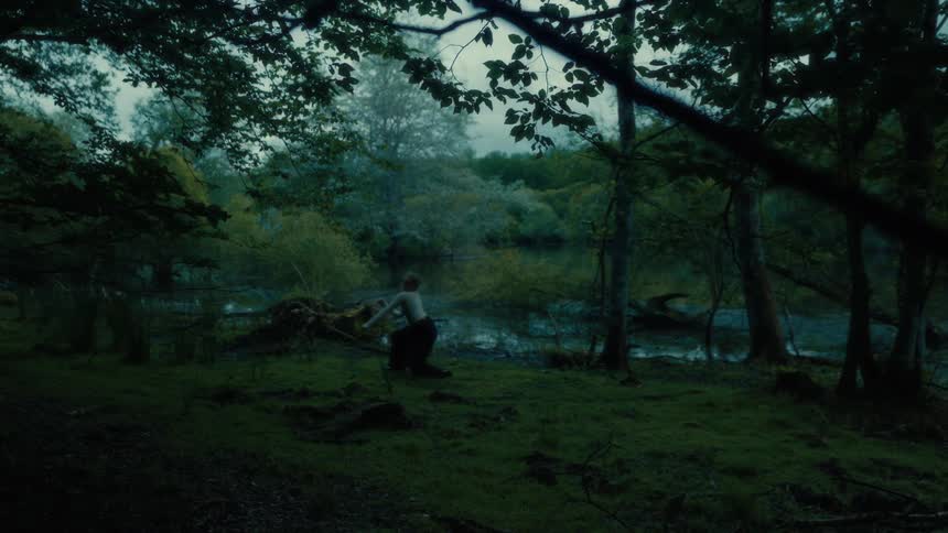 A woman dances slowly in a dimly lit forest near a river