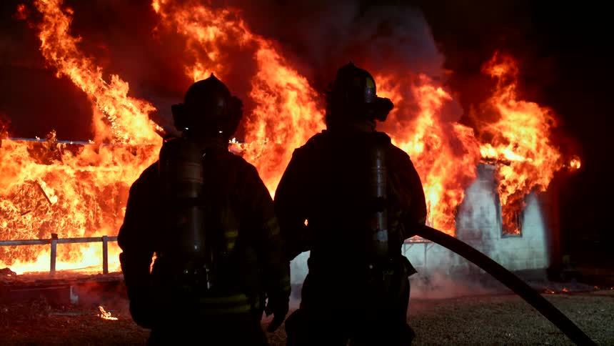 Two firefighters in full gear are silhouetted against a raging fire aiming a hose toward the flames