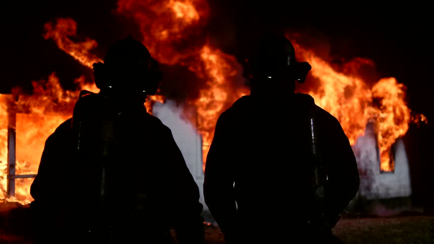 Two firefighters in silhouette stand in front of a large intense fire
