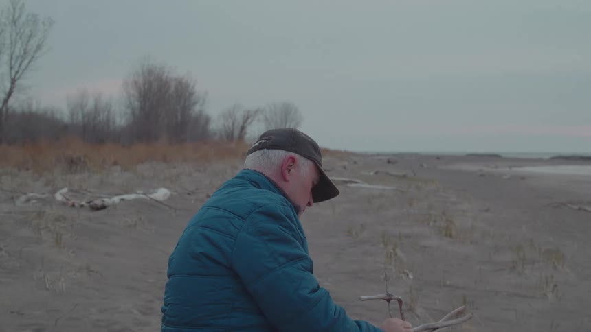 The man holds up a small piece of driftwood on the beach