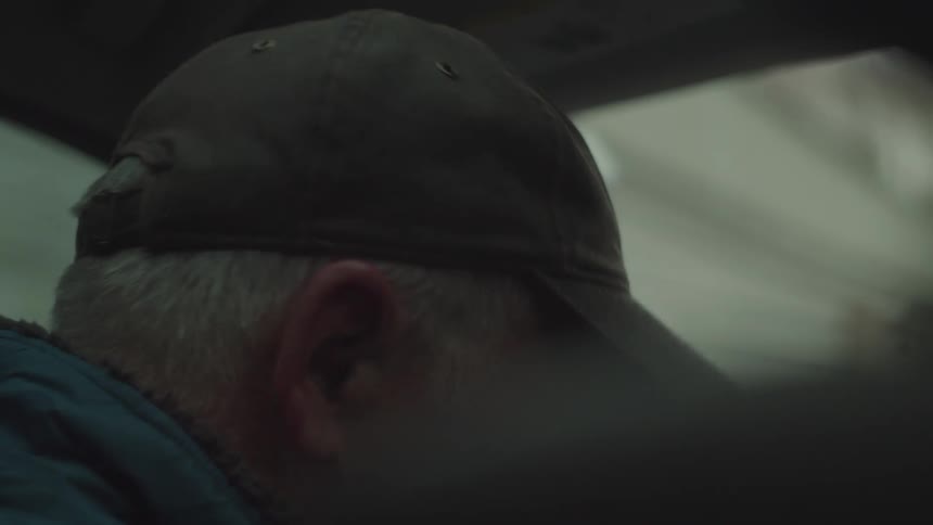 An elderly man wearing a hat and glasses sits behind the steering wheel around midday