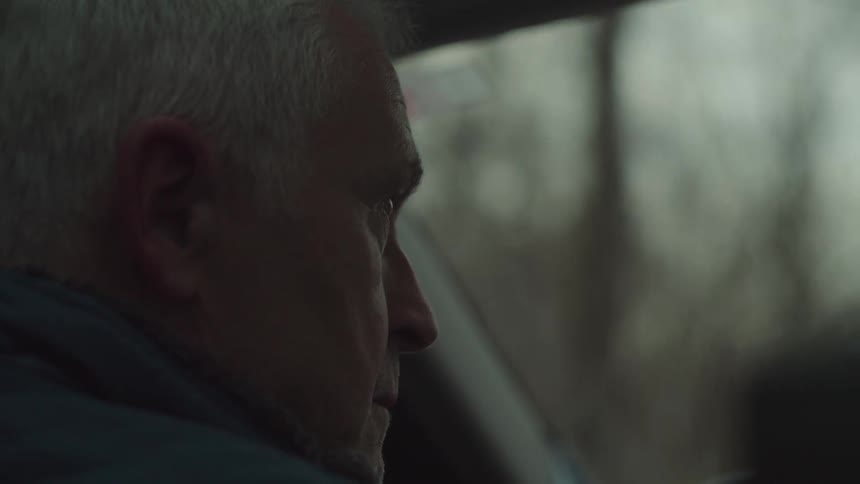 An elderly man drives on the road around midday passing leafless trees