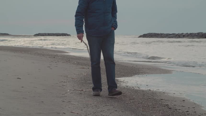 Man walking on the beach poking a stick into the sand as waves roll in to shore