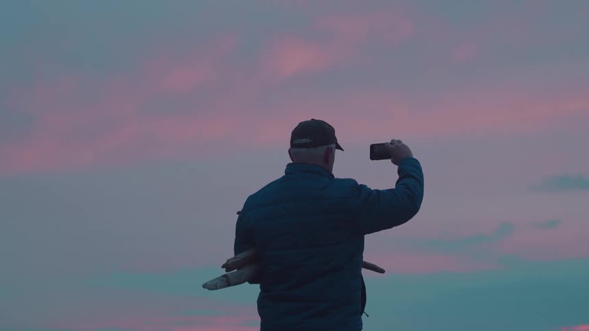 Carrying driftwood in one arm a man in a dark jacket and cap raises his phone to photograph the pink and blue sunset
