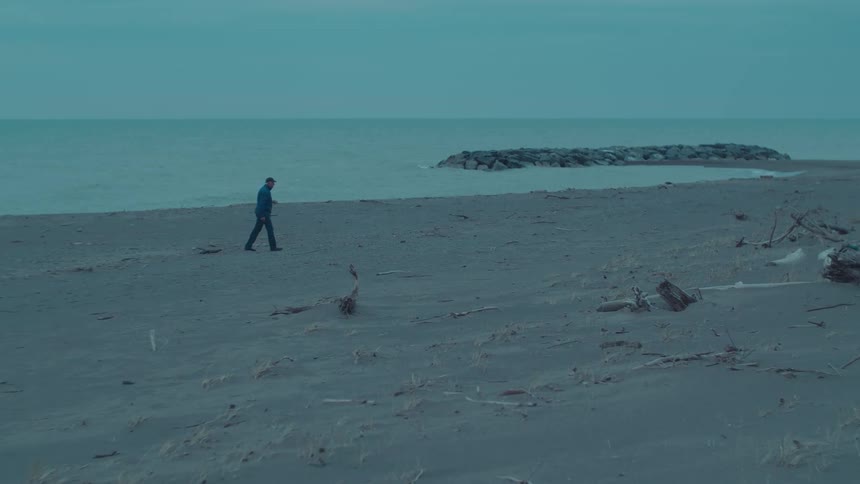 A man walks down the shore of a sandy beach while throwing twigs in the sand