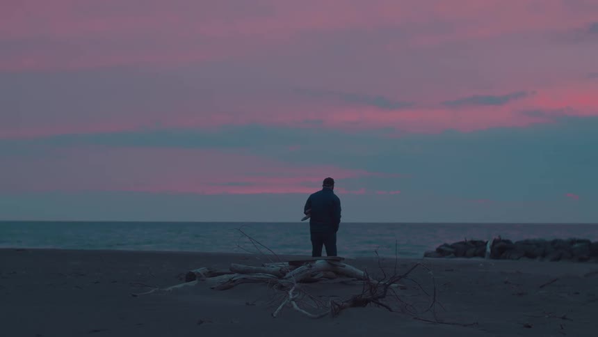A person stands alone on a beach at dusk facing the ocean while collecting driftwood scattered across the sand