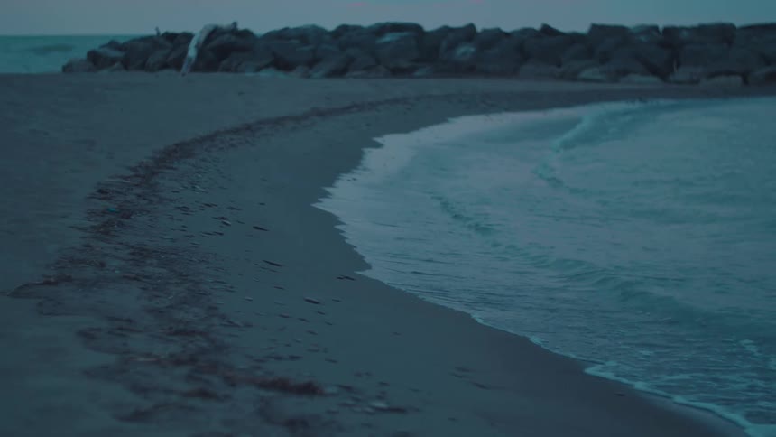 Gentle waves roll onto a quiet beach at dusk with a rocky barrier in the distance