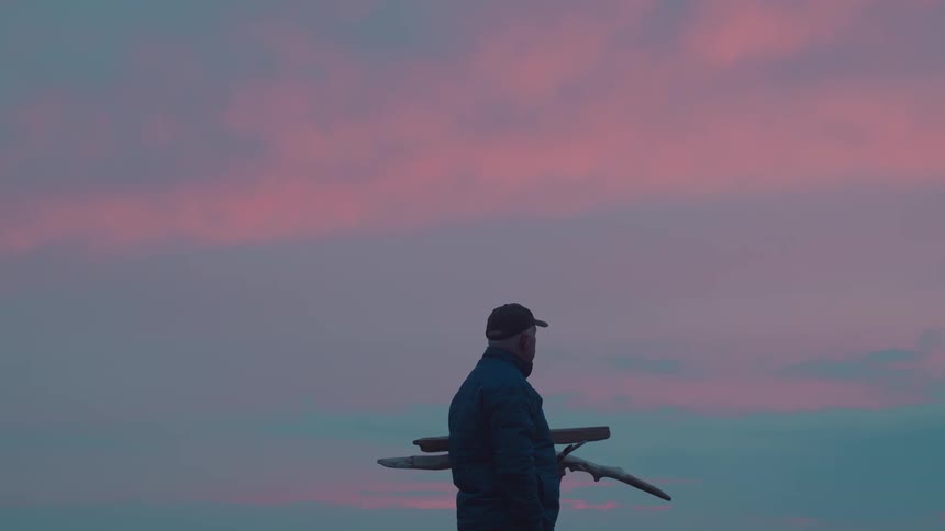 Man in cap in outdoor environment with pink sky holding wooden twigs and logs
