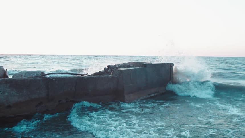 Powerful choppy waves crash against a weathered concrete seawall sending a plume of white spray into the air under a bright sky